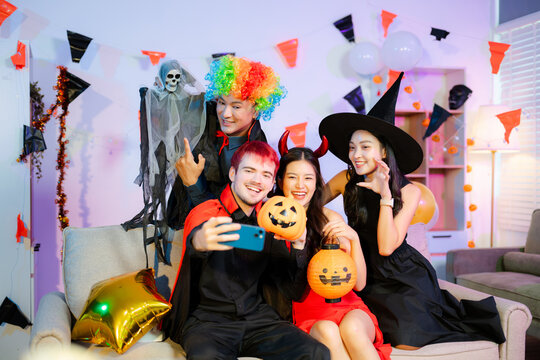 Smiling friends in clown, devil, witch, and vampire costumes take a fun selfie with pumpkins and lanterns at a colorful Halloween party celebration.