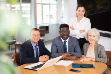 In consulting room of office, positive management team sits at desk and poses for reporting photo. Snapshot of successful businessmen after making profitable deal