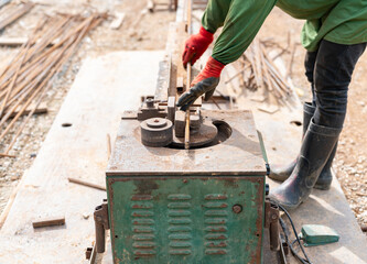 A construction worker, wearing safety gloves and work boots, operates a mechanical rebar bending machine on a building site.