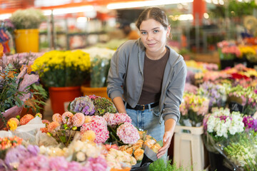 Near showcase with cut flowers, girl visitor examines bunches of hydrangea , selects flowers, and...
