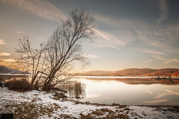 Worthersee lakeshore at dusk in winter near Klagenfurt, Austria. Calm water, light snow on the...