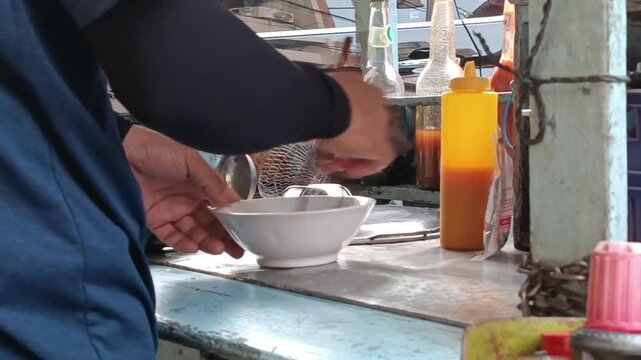 A vendor preparing food at a street food stall. Hands are seen pouring ingredients from a plastic bag into a white bowl. In the background, there are condiment bottles with yellow and red sauces.