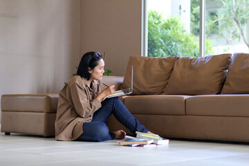 Asian businesswoman sitting on the floor in her living room working with a laptop and notebook with a pen in a serious manner. Workspace