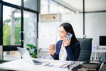 Confident Asian businesswoman using smartphone and drinking coffee at office desk with laptop and documents.