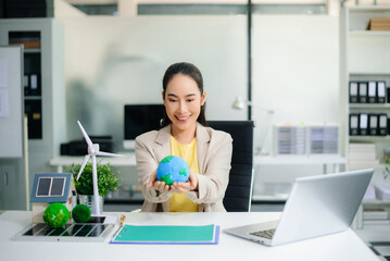 Asian woman in office holding earth model beside solar panels and wind turbine. Concept of sustainability, climate change, and clean energy innovation.