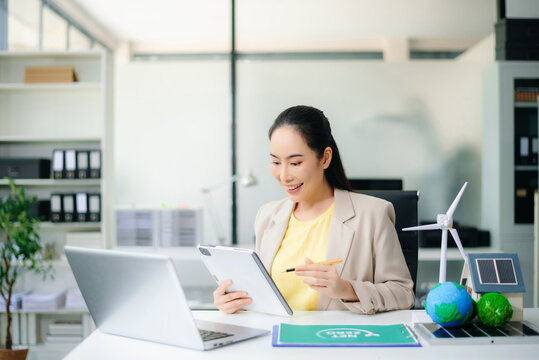 Asian woman holding smartphone and pen near globe, wind turbine, and solar panel on desk. Concept of clean energy, green business, carbon reduction, and smart communication.