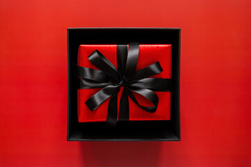 Black Friday concept with an open black box reveals a red gift with a black ribbon on a red background for signaling a secret deal.