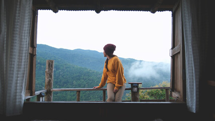 A woman sitting on wooden balcony with mountain views on foggy morning outside the window