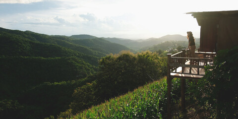A woman standing on a wooden terrace and looking at the forest and mountain views before sunset