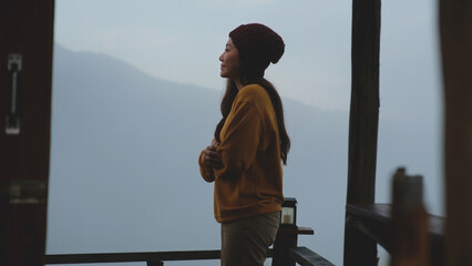A woman standing on wooden balcony with mountain views on foggy morning outside the window