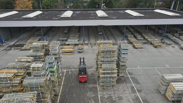 Aerial of a forklift truck driving over an industrial storage yard. The camera tilts up to a large warehouse and reveals high stacks of construction materials