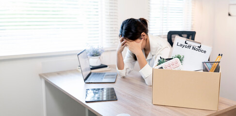 A young Asian businesswoman looking stressed and disappointed after receiving a layoff notice at the office, sitting next to a box of personal belongings.