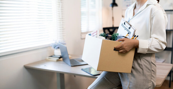 Close-up of an Asian female employee carrying a cardboard box filled with personal items, leaving the office after a layoff or resignation.