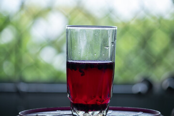 A refreshing glass of red berry juice or fruit tea with fresh berries, sitting by a window with a blurred green garden background.