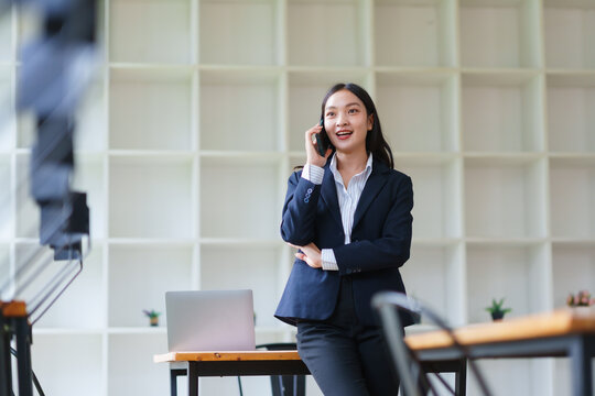 Young Asian businesswoman having a phone conversation in a modern office, standing by a desk with a laptop