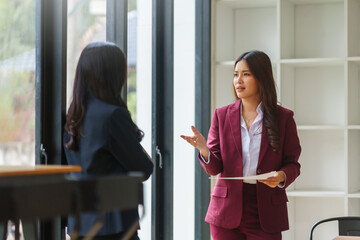 Businesswomen conversing in an office setting. One woman is gesturing while holding documents, discussing with her colleague