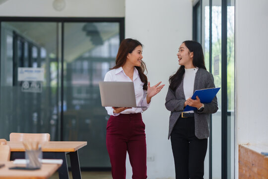 Asian businesswomen discussing work, holding laptop and clipboard, collaborating while walking through a modern workspace - Powered by Adobe