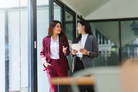 Two Asian businesswomen discussing work while walking outside a modern office building, one holding a digital tablet