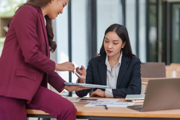 Two Asian businesswomen discussing project data, sharing ideas, and analyzing information on a digital tablet in an office environment