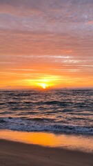 Obraz premium A breathtaking ocean sunset with vivid orange, pink, and purple clouds reflecting on gentle waves. A lone sailboat drifts on the horizon, capturing the serene and dramatic beauty of twilight at sea.