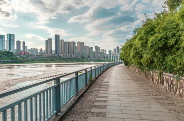 An empty riverside footpath in a green park with the modern city skyline and commercial buildings across the water in Chongqing.