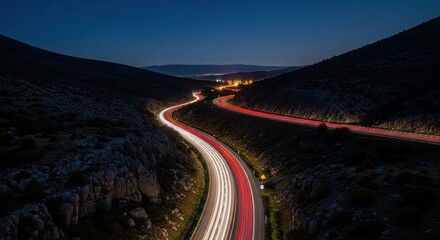 Curved mountain road at night with long exposure car light trails and city glow.