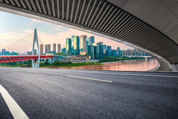 Fototapeta premium Empty asphalt road viewed from under an overpass framing the modern city skyline with a bridge and river at sunset.