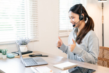 A delighted Asian woman wearing a casual sweater and headset, giving a 'two thumbs up' gesture during a successful virtual meeting or online training session from her home office.