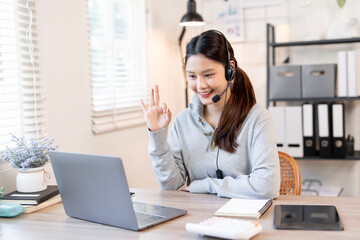 A cheerful Asian woman wearing a casual sweater and headset, giving an 'OK' hand gesture during a virtual meeting or online customer support call from her home office.