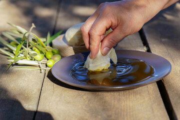 Hand dipping a piece of fresh bread into a shallow plate of extra virgin olive oil, with olives and olive branches on a rustic wooden table