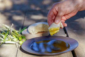 Hand dipping fresh bread into a plate of extra virgin olive oil on a rustic wooden table, with olives and olive branches