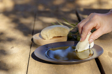 Hand dipping fresh bread into a plate of extra virgin olive oil on a rustic wooden table, with olives and olive branches