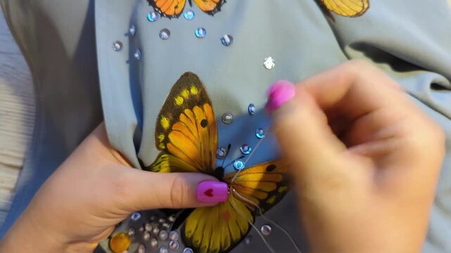 Female hands with pink nails carefully sewing an orange butterfly applique on cloth, demonstrating patience and artistic skill. Hand painted butterfly and beads handiwork.