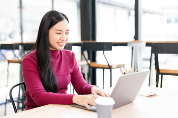 A happy and focused Asian businesswoman smiling while working on her laptop and enjoying a cup of coffee at a modern co-working space.