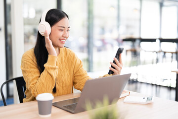 A cheerful Asian woman in a casual yellow sweater wearing headphones, smiling while looking at her mobile phone during a work break or online meeting in a modern workspace.
