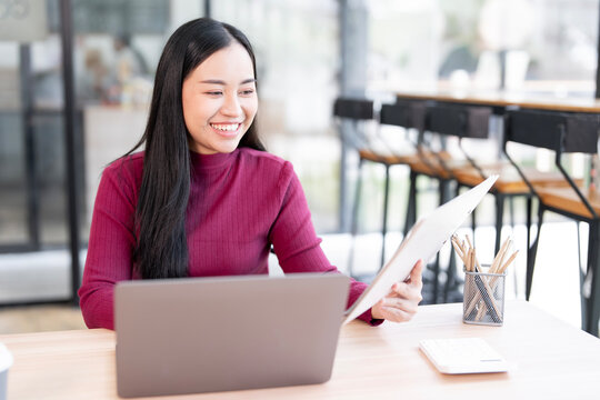 A delighted Asian businesswoman smiling while reviewing documents or reports on a clipboard, working efficiently on her laptop at a modern workspace.