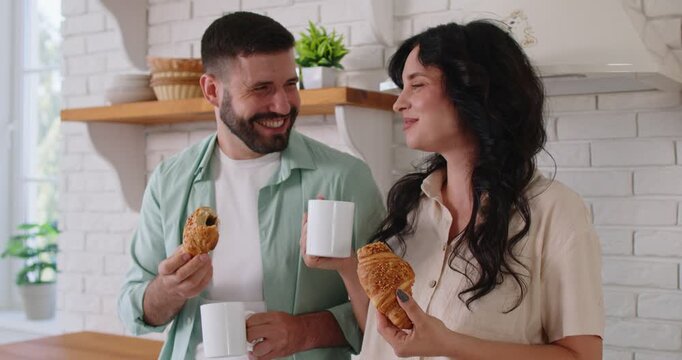 Couple people enjoy breakfast with croissants and coffee in cozy home kitchen. Smiling partners standing together in room, holding mugs and tasty snacks, comfort and affection of morning moment