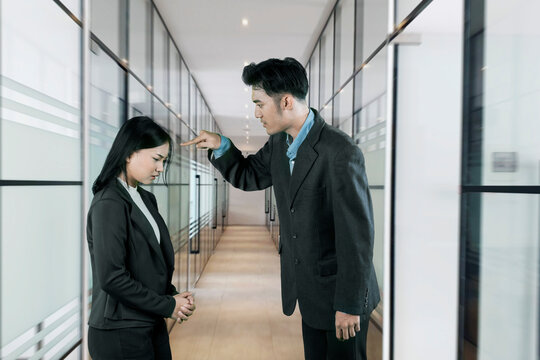 Angry Asian male manager scolding or blaming his sad, submissive female employee in a modern office hallway, depicting workplace conflict and harassment.