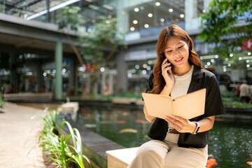 Asian woman multitasking, talking on phone and reading outdoors