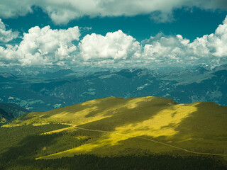 Sunlit mountain slopes in the Dolomites, Italy. Vibrant green meadows and forested hills bathed in golden light beneath dramatic summer clouds — a breathtaking view of alpine beauty.