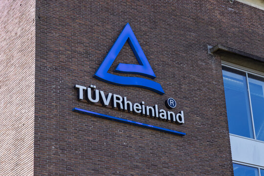 The TUV Rheinland logo sign, a blue triangular symbol and white lettering, mounted on the brick facade of a commercial office building. Arnhem, Netherlands. 19 September 2025.