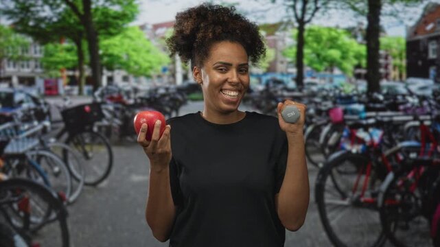 Woman holding red apple and 1kg dumbbell on street in amsterdam wearing black tshirt smiles at camera; healthy fitness joy.