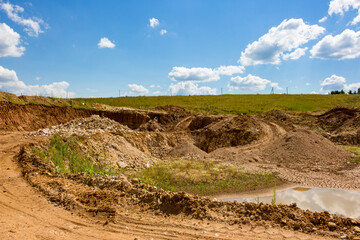 Expansive open-pit quarry under a bright sky, showcasing excavated earth, dirt mounds, and vehicle tracks. A raw, industrial landscape with natural elements