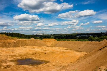 Vast open-pit quarry with rugged, excavated terrain and a murky puddle. Mounds of earth dominate under a bright sky, suggesting ongoing land alteration