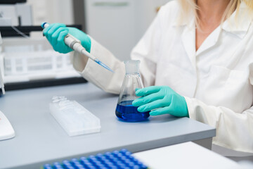 Blonde female scientist in lab carefully using pipette to prepare chemical solution in glass flask....