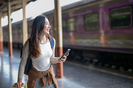 A woman is standing at a train station with a backpack and a cell phone