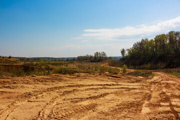 Old sand quarry terrain with numerous vehicle tire tracks. Barren slopes and distant green trees under a bright blue sky, revealing a rugged outdoor landscape