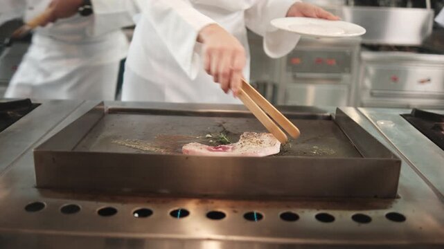 Close-up shot of chef hand-roasting a beef steak with tongs in frying pan, herbs and spices cooking ingredients in restaurant kitchen, professional in gourmet foods and delicious meal occupation.  