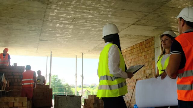 Engineers with blueprint discuss progress while builders bricklaying on construction site. Safety inspectors control incomplete building condition