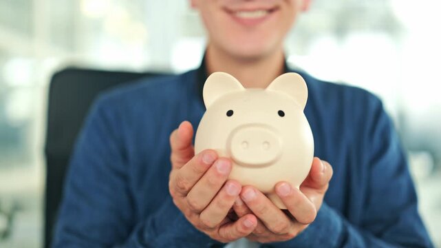 Cheerful man holds a beige piggy bank in an office, wearing a blue shirt. Smiling male embodies the concept of financial planning, savings, and future investments with happiness.
